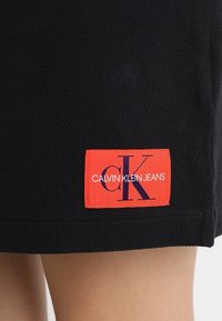 Černá texturovaná látka s červeným obdélníkovým štítkem s nápisem "CK Calvin Klein Jeans" blízko lemu nad odkrytou pokožkou.