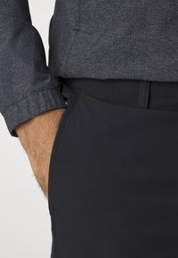 Manche de chemise grise texturée à côté d'un pantalon noir à la finition lisse ; le pantalon est doté de poches latérales et d'une ceinture plate.