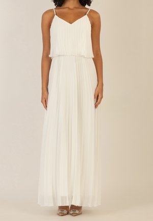 Femme portant une robe maxi plissée blanche à fines bretelles et des escarpins à bout pointu ornés de cristaux, posant devant un fond uni.