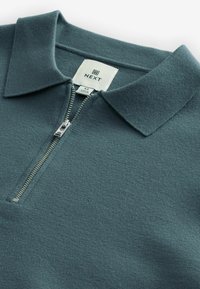 Zelenomodrá polo košile na zip vyrobená z texturované tkaniny. Obsahuje krátký límec a stříbrný zip s označením značky uvnitř.