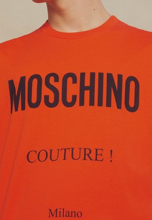 Osoba nosząca jasnopomarańczową koszulkę z dużym czarnym tekstem "MOSCHINO" oraz mniejszymi tekstami "COUTURE!" i "Milano" poniżej.