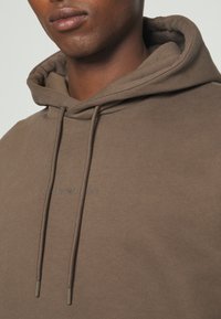 Brown hooded sweatshirt made of smooth cotton fabric. Features a kangaroo pocket and adjustable drawstrings. Logo printed on the chest.