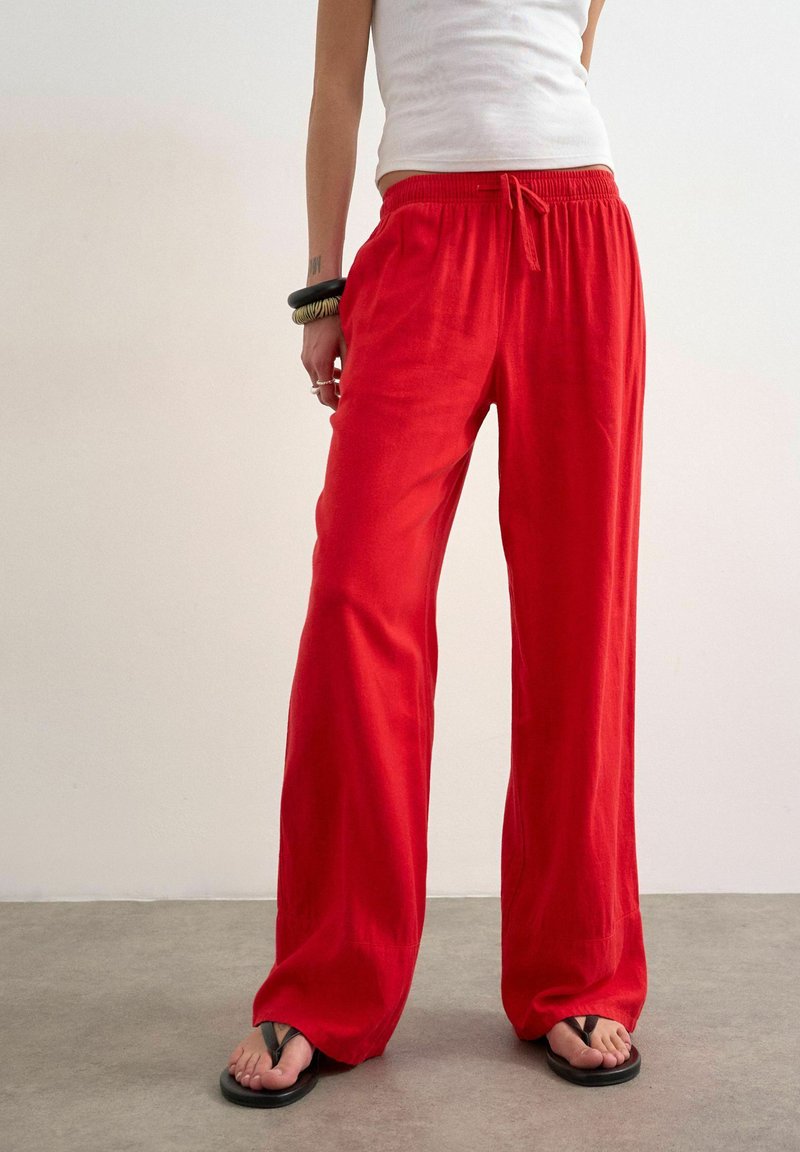 Personne portant un pantalon rouge ample avec cordon, un haut blanc sans manches, des tongs noires et des bracelets, debout sur un sol gris.