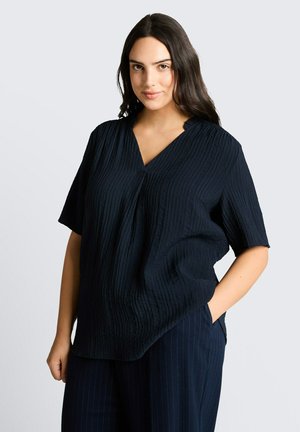 Jeune femme aux longs cheveux foncés portant un haut bleu marine texturé à col en V et manches courtes, et un pantalon bleu marine à fines rayures, posant avec une main dans la poche.