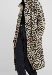 Płaszcz w panterkę z miękkiego materiału w szarym odcieniu, z czarnymi i brązowymi plamami, bocznymi kieszeniami oraz luźnym fasonem. Zestawiony z ciemnymi dżinsami.
