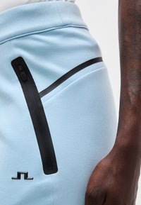 Prim-plan cu pantaloni de culoare albastru deschis, cu buzunar cu fermoar negru și logo negru în apropierea șoldului, alături de un braț cu piele închisă la culoare odihnindu-se pe lângă corp.