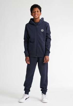 Felpa con zip blu navy in cotone, dotata di cappuccio, tasche frontali e un logo circolare, abbinata a pantaloni jogger coordinati e sneaker bianche.