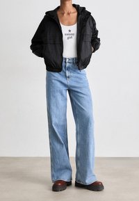 Svart lättviktsjacka med huva, två framfickor; vit linne med textlogotyp; ljusblå vida jeans; bruna platåskor.