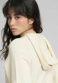 Sudadera con capucha de color crema y textura suave, con un ajuste relajado y capucha caída. Cabello y rostro parcialmente visibles, con fondo neutro.