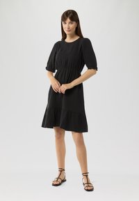 Schwarz, knielanges Kleid mit tailliertem Schnitt, puffähnlichen Ärmeln und gestuften Saum. Besitzt eine glatte Textur und ein einfaches Design. Gestylt mit schwarzen Sandalen.