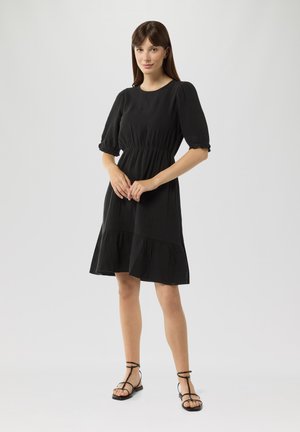 Schwarz, knielanges Kleid mit tailliertem Schnitt, puffähnlichen Ärmeln und gestuften Saum. Besitzt eine glatte Textur und ein einfaches Design. Gestylt mit schwarzen Sandalen.