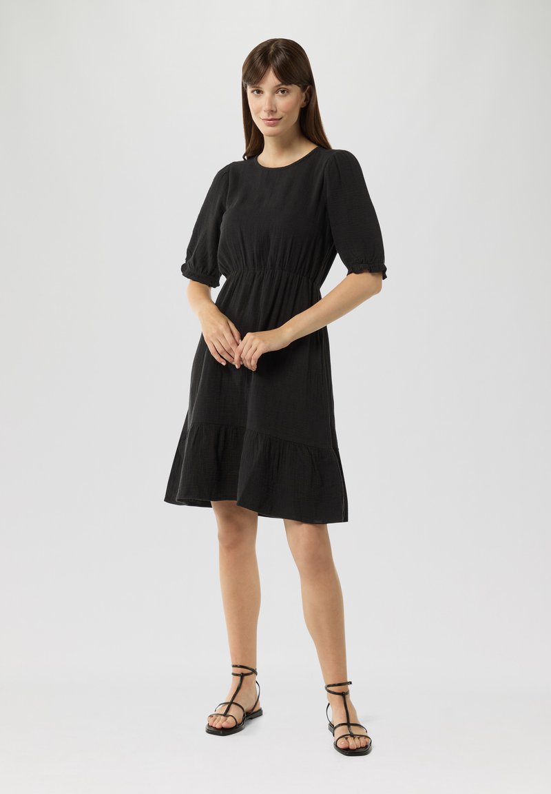 Schwarz, knielanges Kleid mit tailliertem Schnitt, puffähnlichen Ärmeln und gestuften Saum. Besitzt eine glatte Textur und ein einfaches Design. Gestylt mit schwarzen Sandalen.