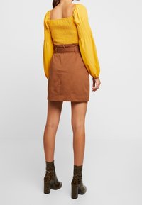 Top giallo con arricciature e maniche a sbuffo, abbinato a una mini gonna marrone. La modella indossa stivali alla caviglia color oliva con stampa coccodrillo e tacco a blocco.