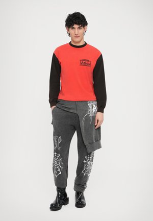 Red and black long-sleeve shirt with logo; grey sweatpants featuring white spider web and graphic designs; black shiny boots.