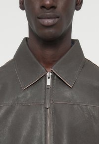 Γκρι δερμάτινο μπουφάν με μυτερό γιακά, διαθέτει κλείσιμο με φερμουάρ μπροστά και ορατές ραφές σε πιο ανοιχτή απόχρωση.