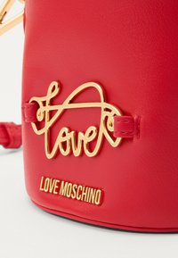Punainen tekonahkainen käsilaukku, jossa on kultainen "love"-logo ja kultainen "LOVE MOSCHINO"-symboli. Sileä pinta ja kompakti muotoilu.