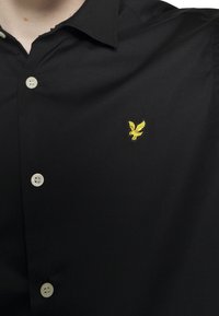 Camicia nera con bottoni, dotata di un piccolo ricamo di un uccellino giallo sul lato sinistro del petto.