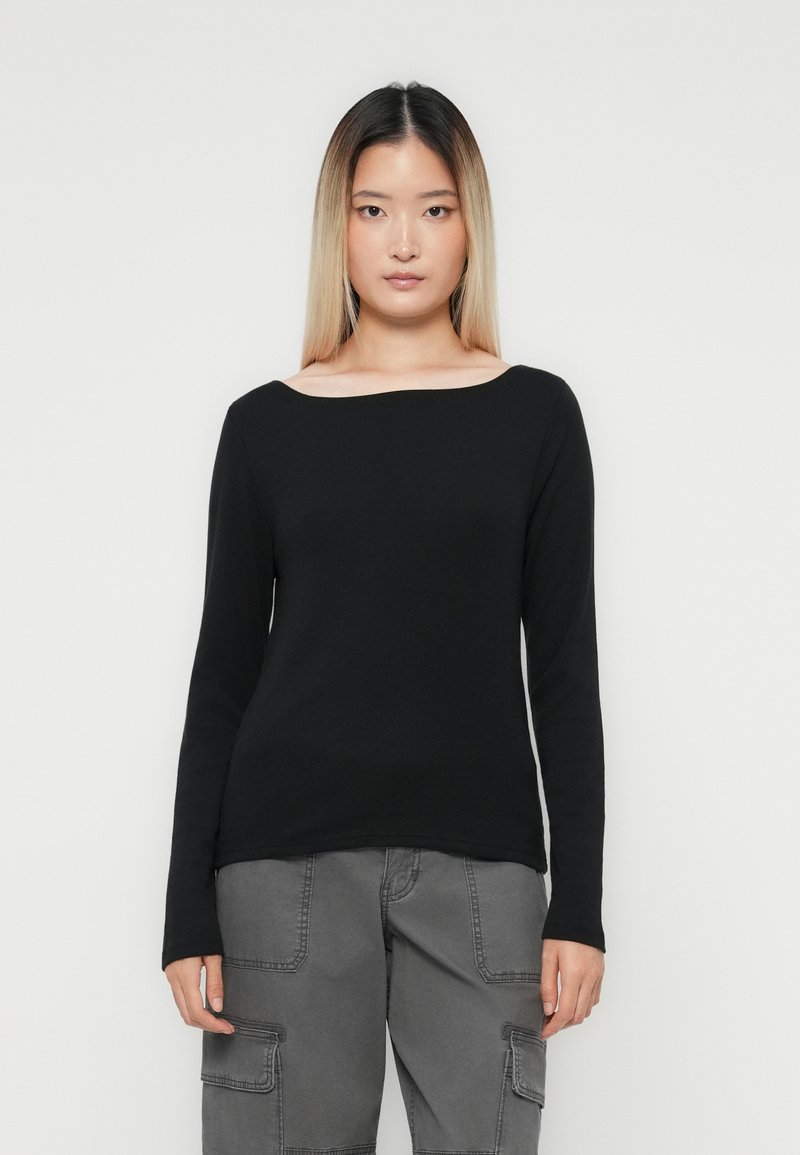 Een jonge vrouw met stijlvol blond haar draagt een zwart shirt met lange mouwen en grijze cargobroek, terwijl ze tegen een effen witte achtergrond staat.