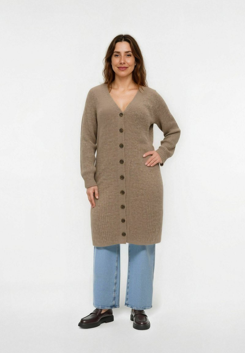 Beige Strickjacke mit V-Ausschnitt, geknöpftem Vorderteil und geripptem Muster. Kombiniert mit weit geschnittenen blauen Jeans und schwarzen Loafers.