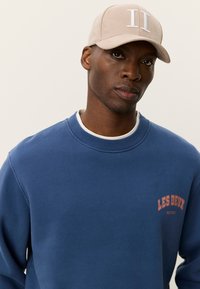 Μπλε σκούρο φούτερ με ριμπ μανσέτες και στρόγγυλη λαιμόκοψη, με κόκκινο λογότυπο «LES DEUX». Μπεζ καπέλο με λευκή κεντημένη επιγραφή «II» στο μπροστινό μέρος.