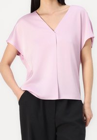 Blusa corta rosa chiaro con maniche corte e scollatura a V, con dettaglio plissettato sul davanti e vestibilità rilassata, realizzata in tessuto morbido.