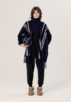 Marine poncho met grijze strepen en gepofte randen, gedragen over een zwarte coltrui en broek, gecombineerd met een zwarte handtas en bruin, met bont gevoerde laarzen.