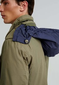 Olive green jacket with a visible navy hood, zippered detailing on the collar, and textured fabric. Features a button on the hood attachment.