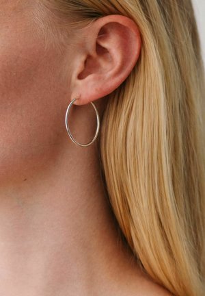 Približni posnetek osebe z blond lasmi, ki nosi tančico srebrno obrobo na levem ušesu, prikazuje teksturo kože in pramene las.