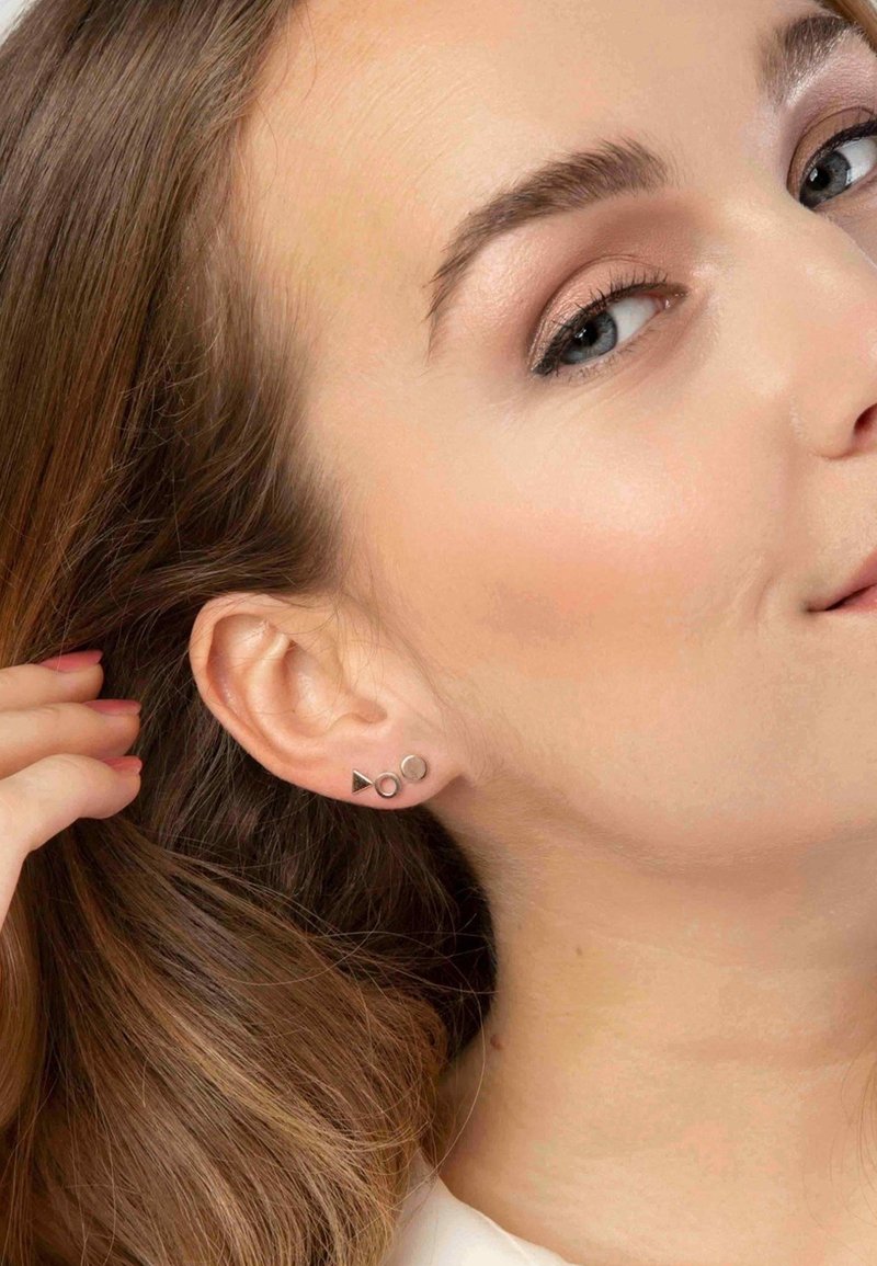 Zilveren studs met driehoekige, ronde en ovale vormen, gedragen in het oor van een model met lichtbruin haar dat gedeeltelijk bedekt is.