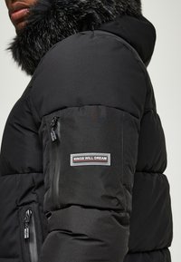 Schwarze Latzjacke mit einer abnehmbaren Kapuze aus Kunstpelz. Verfügt über eine Reißverschlusstasche am linken Arm und einen Logopatch an der Vorderseite.