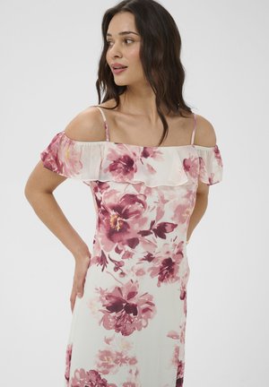 Donna con capelli scuri che indossa un vestito bianco a spalle scoperte con stampa floreale rosa e viola, in piedi con le mani sui fianchi, che guarda verso sinistra.