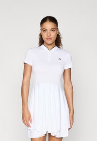 Biała sportowa sukienka z zamkiem błyskawicznym przy kołnierzu, krótkimi rękawami i wielowarstwowym dołem. Posiada małe czarne logo na piersi oraz gładką teksturę.