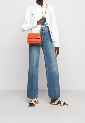 Persoon draagt een witte spijkerjack, blauwe jeans met wijde pijpen, witte sandalen en houdt een oranje crossbodytas met een gouden kettingband vast.