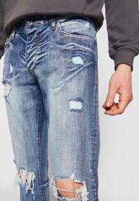 Jean en denim bleu clair avec un design usé, des ourlets effilochés et de grandes déchirures sur les deux jambes. Style authentique à cinq poches.