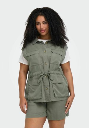 Kvinde med krøllet hår iført en olivengrøn vest med knapper og lommer samt matchende shorts over en hvid T-shirt med korte ærmer.