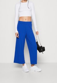 Pantalons bleu à jambes larges avec texture côtelée verticale, associés à un crop top blanc et des baskets blanches. Un petit sac à main noir est tenu dans une main.