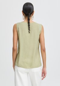 Top senza maniche di un verde oliva chiaro con una texture liscia e orlo arrotondato, abbinato a pantaloni bianchi. Design semplice, senza motivi o accenti visibili.