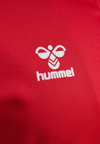 Rode stof met een gladde textuur, met een wit logo van een gestileerde bij en de merknaam "hummel" eronder.