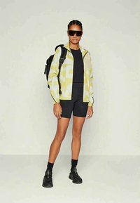 Vrouw in zwarte fietsbroek, zwart topje, gele-witte geprinte jas, zwarte sneakers, zonnebril en zwarte rugzak die tegen een effen achtergrond staat.