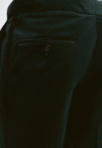 Κοντινή λήψη μαύρου ραμμένου παντελονιού που δείχνει μια πίσω τσέπη με κουμπί στη δεξιά πλευρά.