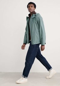 Giacca impermeabile verde con cappuccio, zip frontale e tasca sul petto, indossata sopra una camicia marrone. Abbinata a jeans blu scuro e sneaker bianche.