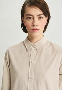 Chemise beige clair en tissu lisse, dotée d'un col traditionnel, d'une fermeture à boutons et de manches longues avec un léger éclat.