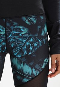 Czarne legginsy z zielonym tropikalnym wzorem liści, wykonane z elastycznej tkaniny z siateczkowymi detalami po bokach dla lepszej wentylacji.