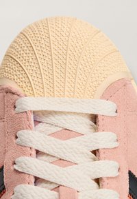 Różowa cholewka z zamszu z białymi sznurowadłami, wyposażona w kremową, teksturowaną gumową podeszwę oraz wzorzysty nosek. Czarny detal z logo.