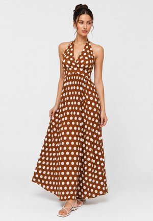 Woman wearing a brown halter-neck maxi dress with white polka dots and white strappy sandals, standing against a plain background.
