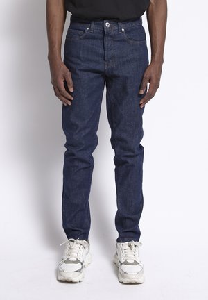 Man in donkerblauwe spijkerbroek en witte sneakers, staand met ontspannen handen langs het lichaam tegen een effen achtergrond.