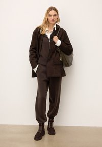 Veste en daim marron à double boutonnage portée sur un pull en tricot marron foncé et un pantalon de jogging assorti. Bottes à lacets marron à la cheville et sac à main olive.