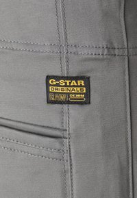 G-Star Originals siva denimska tkanina s vidnim šivi. Oznake vsebujejo črno ozadje z rumenim besedilom: "G-Star," "ORIGINALS" in "RAW DENIM."