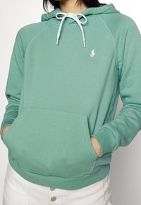 Sudadera verde menta con cordones blancos, bolsillo canguro y puños acanalados. Presenta un logo en el pecho y una textura suave.