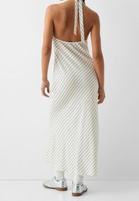 Vit ärmlös klänning med diagonala ränder, öppen rygg, knytkrage och en flödande siluett, ihopparad med silverfärgade sneakers och vita strumpor.